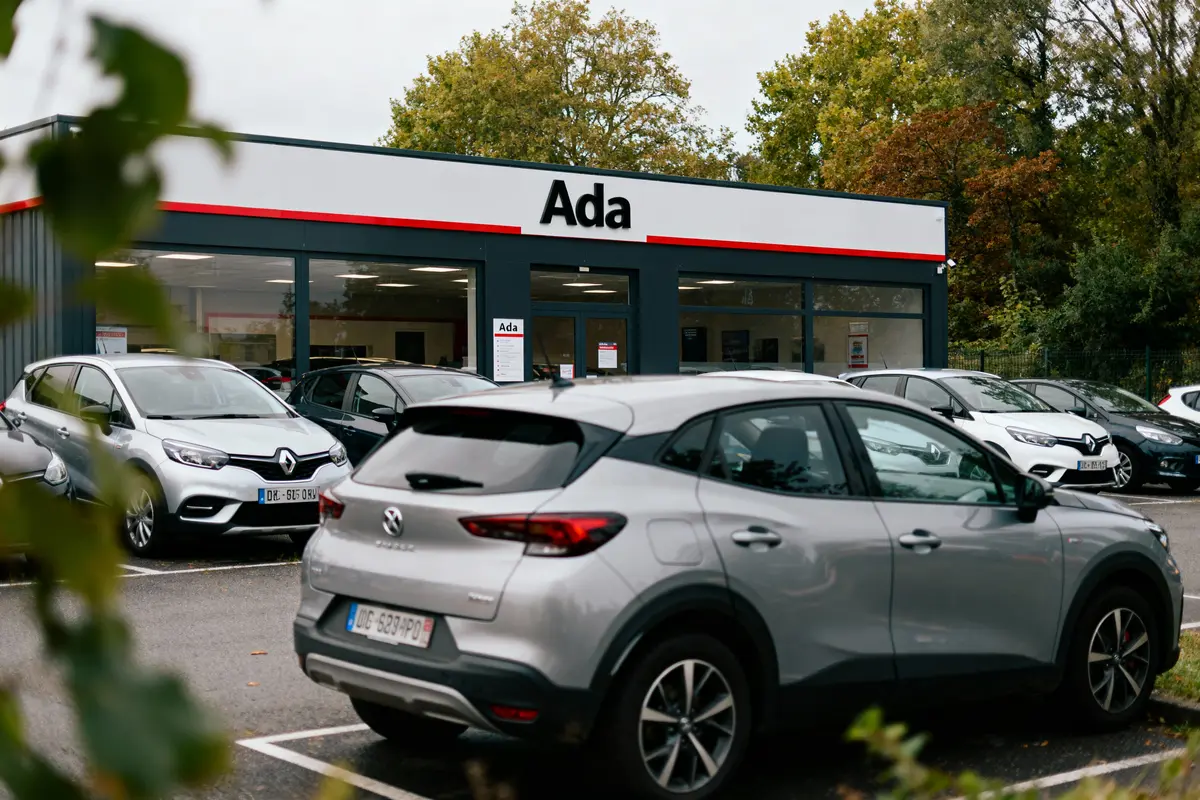 Parking auto devant agence de location Ada, avec plusieurs voitures garées, arbres en arrière-plan.