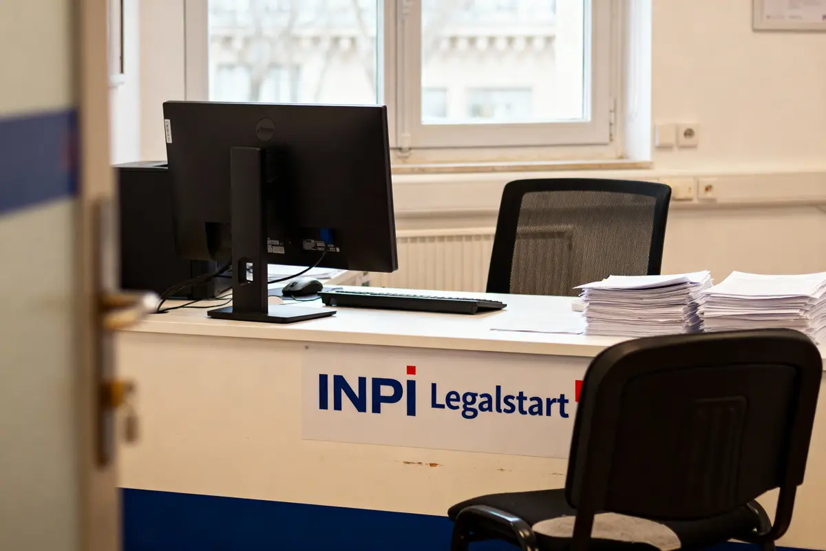 Bureau avec ordinateur, piles de documents, affichage "INPI Legalstart", fenêtre en arrière-plan, chaise noire.