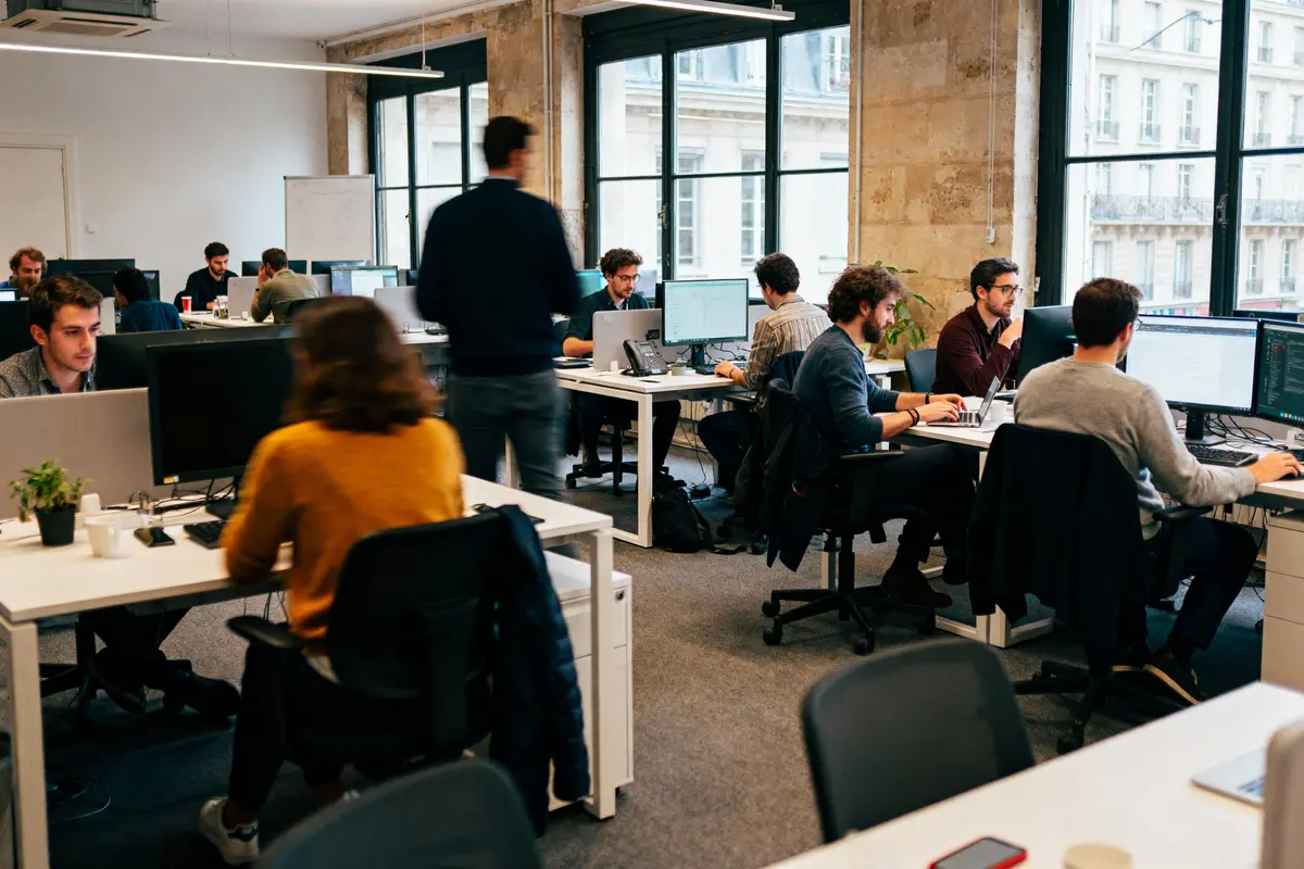 Personnes travaillant dans un bureau moderne, ouvert, avec ordinateurs et fenêtres donnant sur un bâtiment urbain.