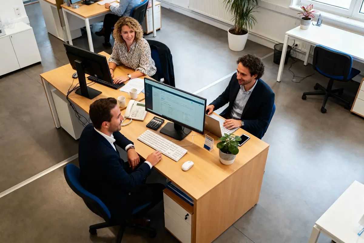 Trois personnes discutent autour d'un bureau partagé, travaillant sur des ordinateurs dans un espace de bureau lumineux.