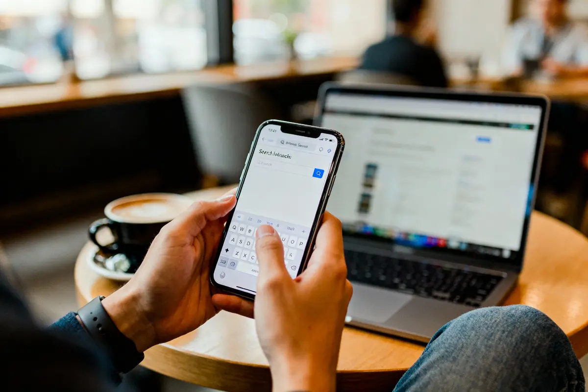 Personne utilisant un smartphone devant un ordinateur portable dans un café, avec une tasse de café.