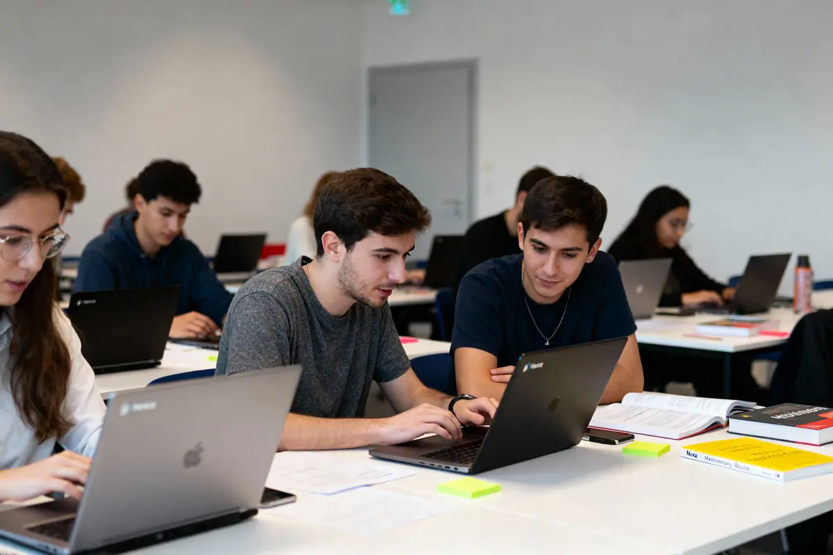 Étudiants travaillant ensemble sur des ordinateurs portables en classe, entourés de livres et de documents.