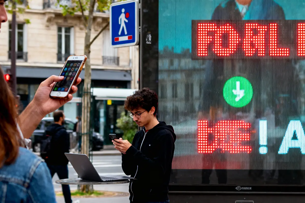 Personnes utilisant smartphones et ordinateur portable devant panneau LED, rue animée avec signalisation piétonne.