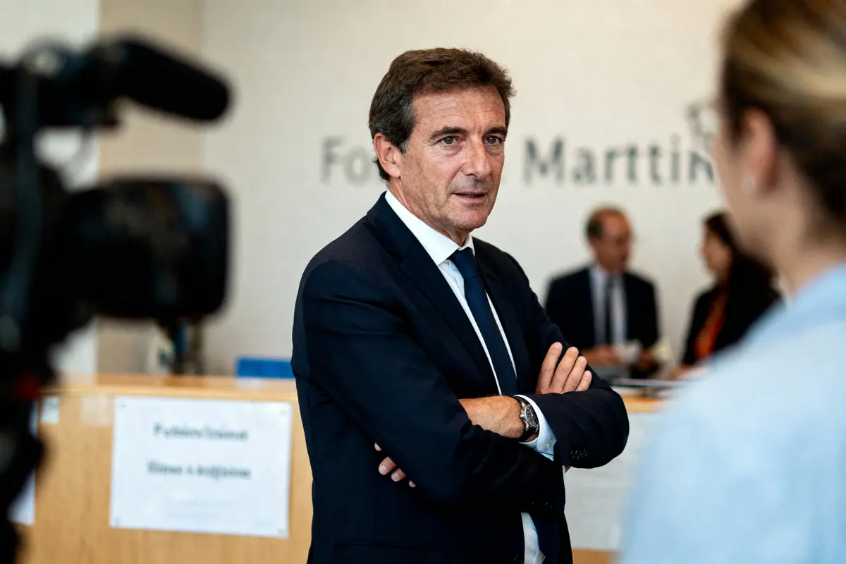 Un homme en costume discute dans un environnement professionnel, avec caméra et personnes en arrière-plan floutées.