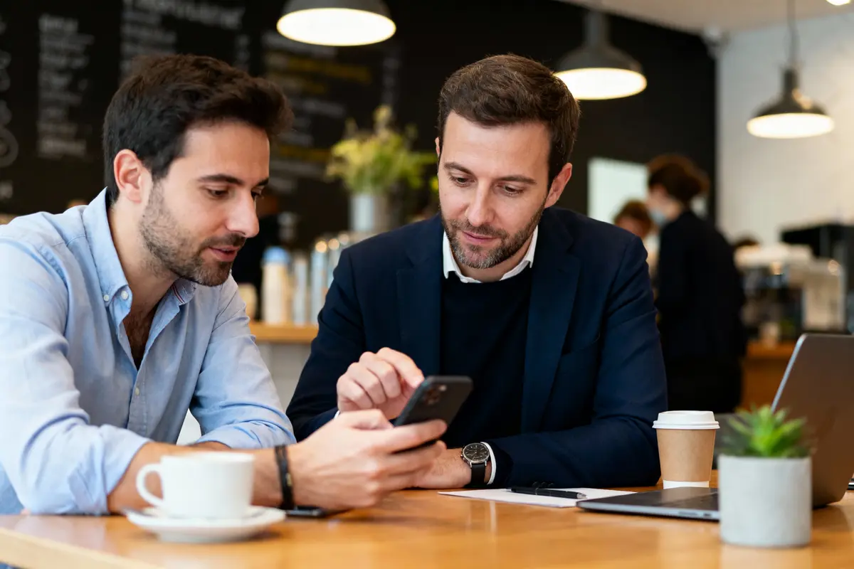 Deux hommes discutent autour d'un smartphone dans un café, devant un ordinateur portable et des tasses.