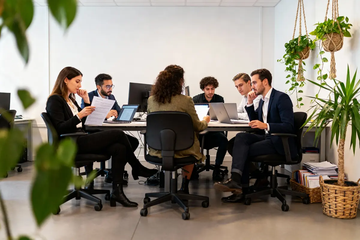 Personnes en réunion autour d'une table avec ordinateurs et documents, plantes suspendues en arrière-plan.