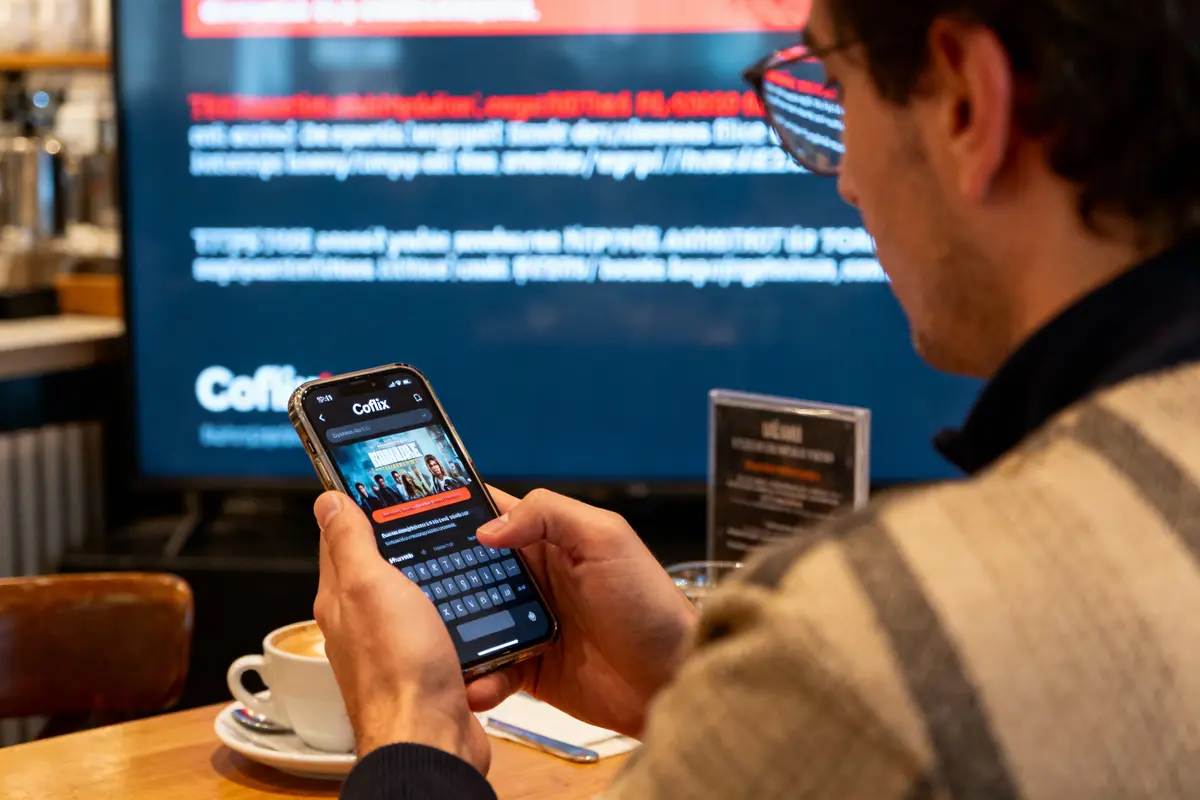 Homme utilisant smartphone dans un café, écran de télévision flou en arrière-plan, tasse de café sur la table.