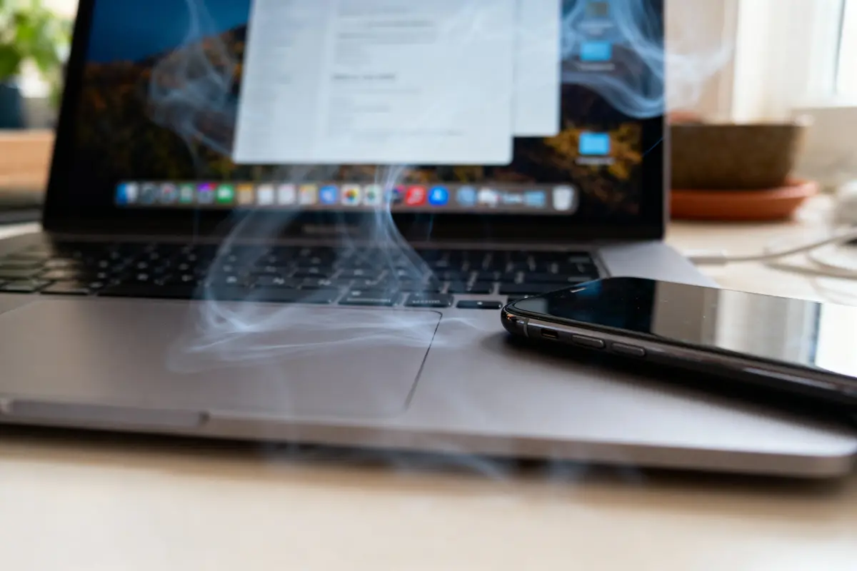 Ordinateur portable avec fumée, écran allumé, smartphone posé dessus, ambiance de surchauffe.