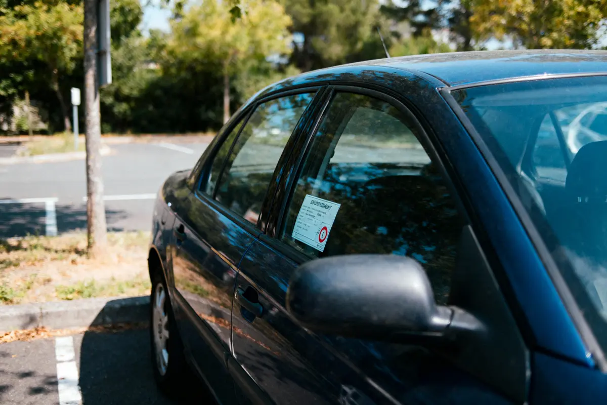 Voiture bleue garée, vitre avec vignette, environnement verdoyant et ensoleillé.