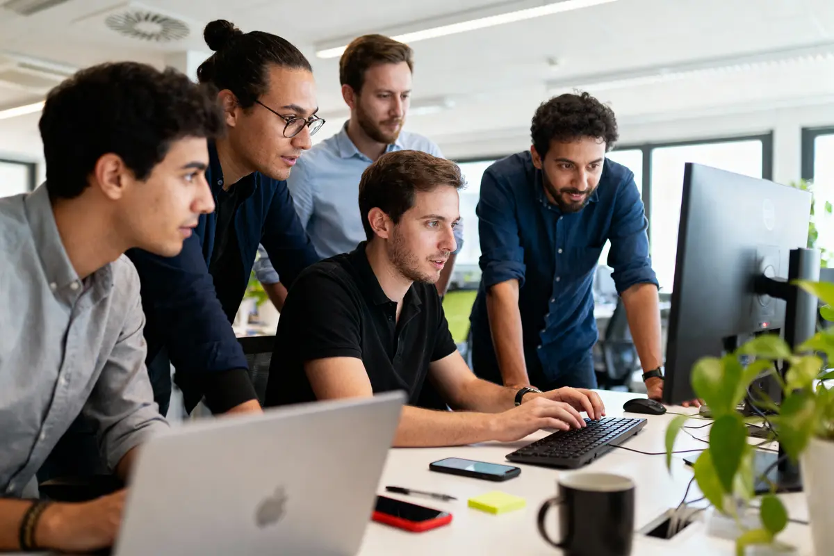 Cinq personnes collaborent autour d'un écran d'ordinateur dans un bureau moderne, travaillant ensemble.