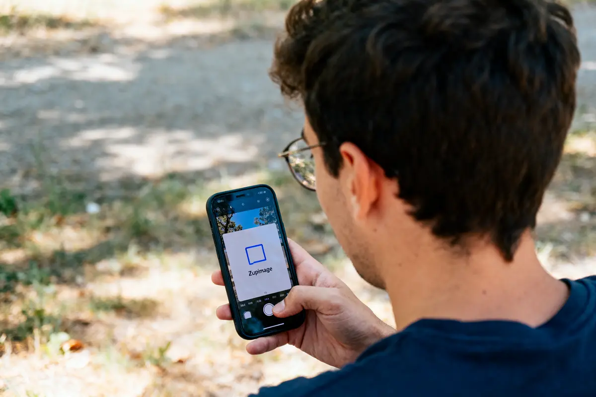 Homme utilisant un smartphone avec application "Zupimage", assis à l'extérieur, arrière-plan flou.