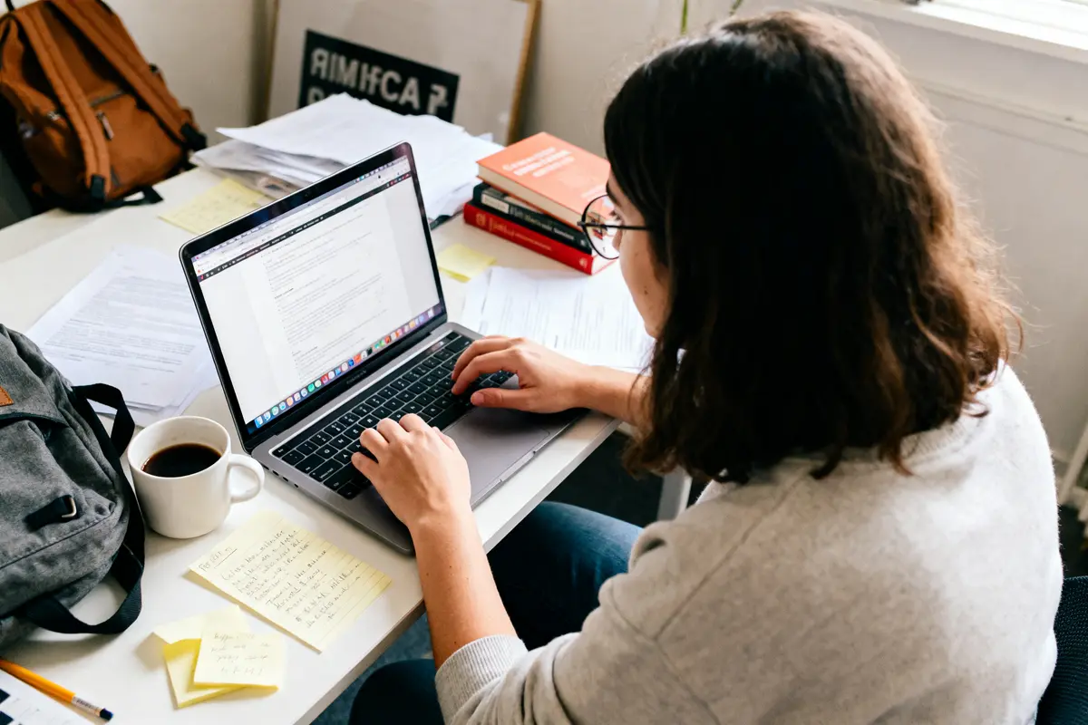 Personne tapant sur un ordinateur portable, bureau encombré, tasse de café, livres, notes et sac à dos.